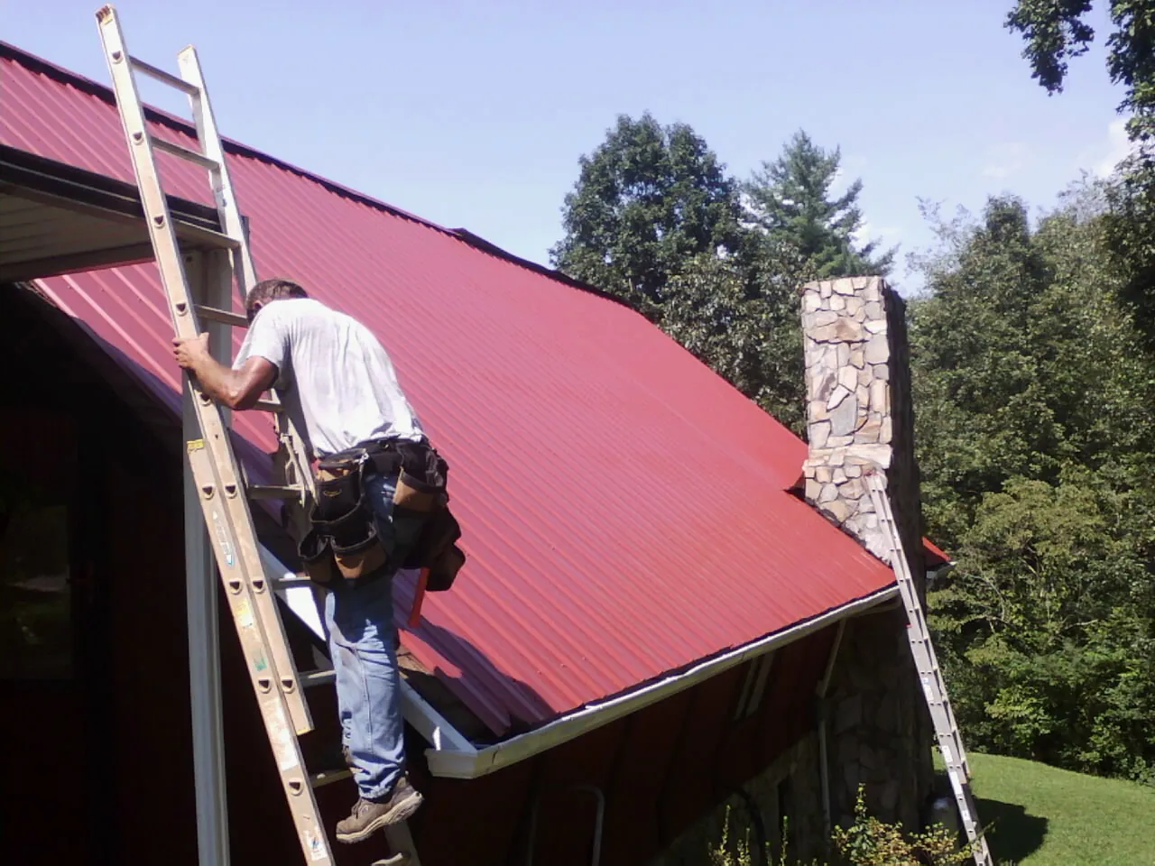 Roofer on a ladder beside a red metal roof and stone chimney — Soffit & Fascia Repair by Roofing Brothers in Creedmoor